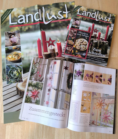 Landlust November/Dezember 2026: Titelseite „Zum Lichterfest“ mit roten Kerzen, Beilage „Plätzchen zum Advent“ und aufgeschlagene Seite mit Papiersternen
